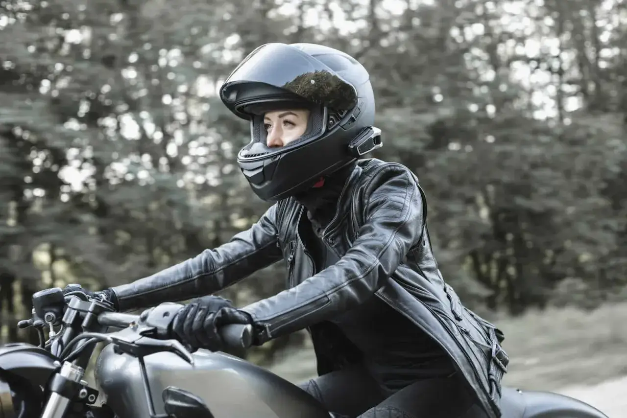 Kobieta w sk&oacute;rzanej kurtce i kasku szczękowym lub integralnym jedzie motocyklem przez las.