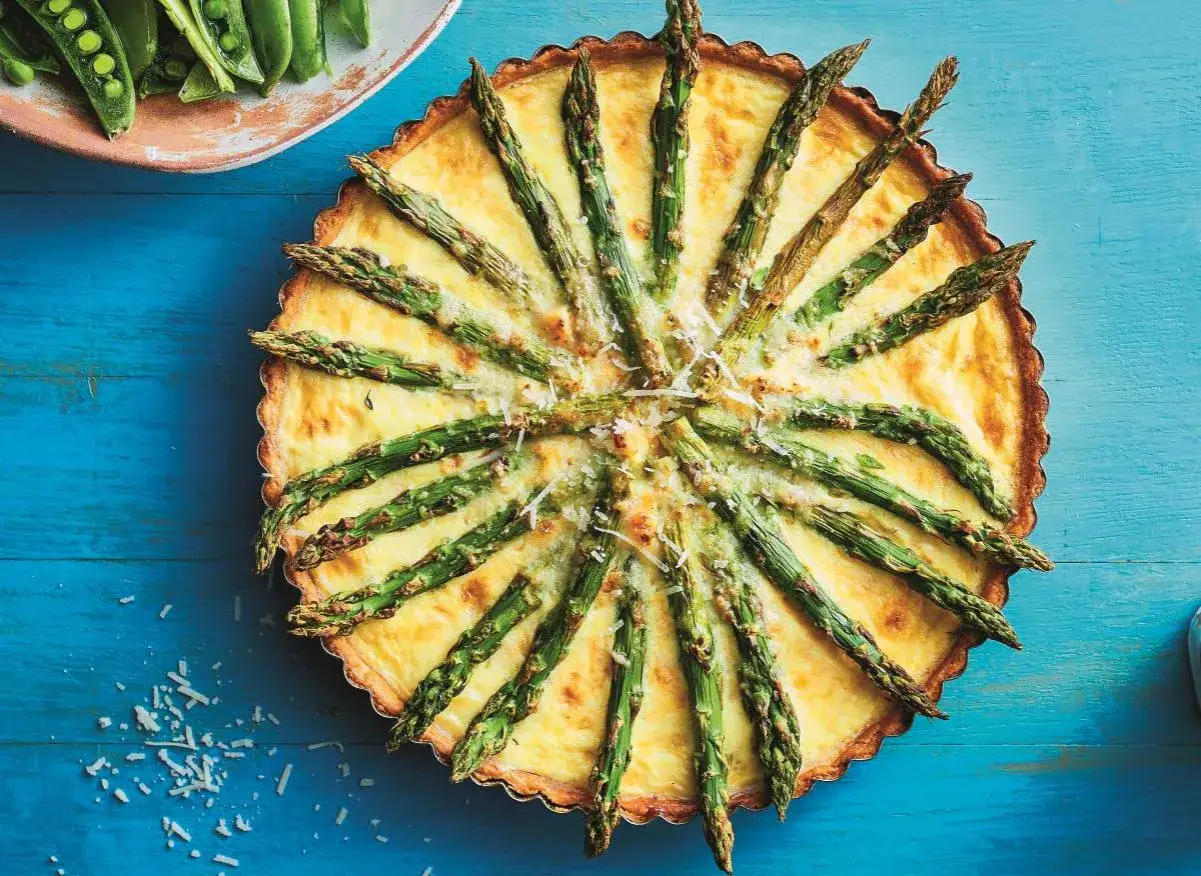 Tarta ze szparagami ułożonymi promieniście na ż&oacute;łtym, kremowym nadzieniu, posypana startym serem. Obok talerz ze świeżymi strąkami groszku.