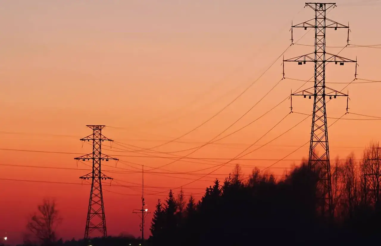 Zachód słońca nad liniami energetycznymi. W tle pomarańczowe niebo, a na pierwszym planie sylwetki słupów wysokiego napięcia i drzew. Warto pamiętać o dopłatach do prądu.