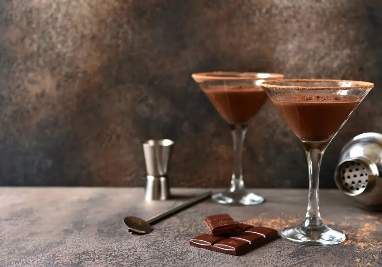 Dwa kieliszki z czekoladowym likierem, posypane kakao. Obok kostki czekolady, shaker i miarka. Idealny przepis na likier czekoladowy.