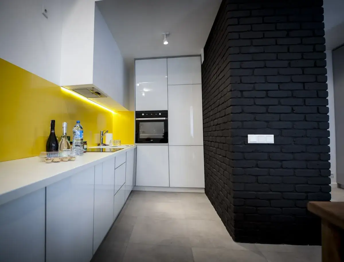 Nowoczesna kuchnia z ż&oacute;łtym panelem i czarną cegłą na ścianie. Białe szafki i blat, wbudowany piekarnik.