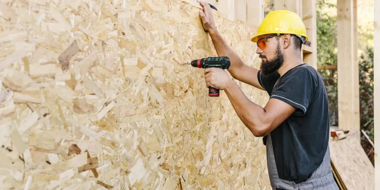 Budowlaniec w żółtym kasku przykręca płytę OSB. Różne rodzaje płyt OSB są kluczowe w budownictwie.