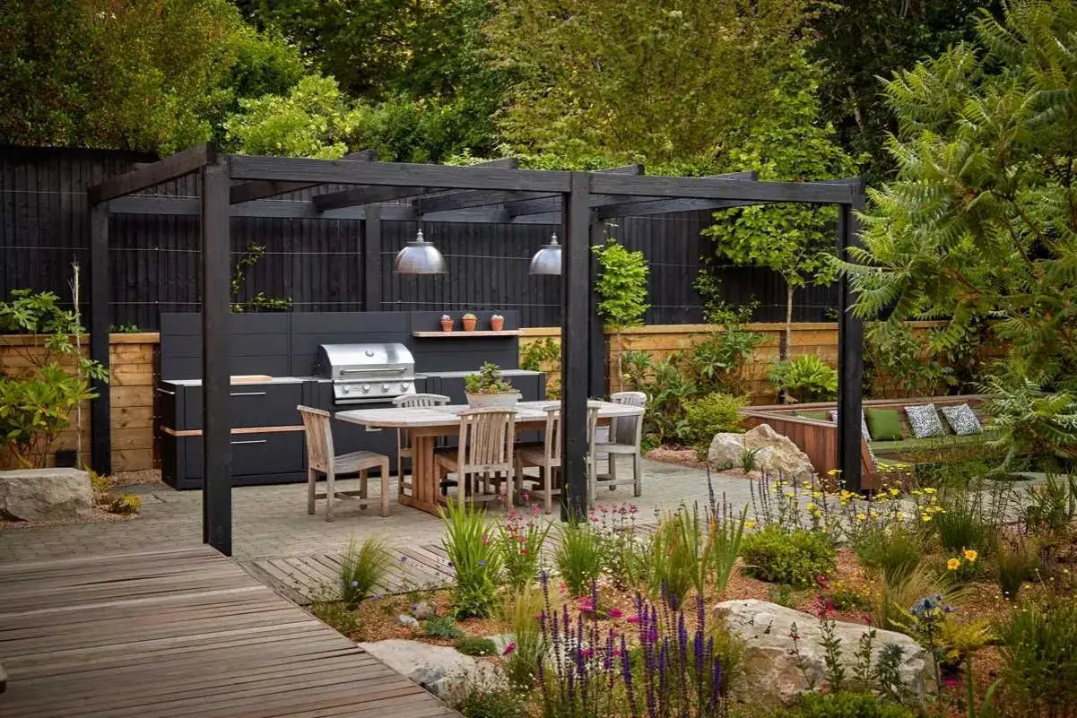 Nowoczesny grill w ogrodzie pod pergolą, obok jadalnianego stołu i wypoczynkowej ławy.