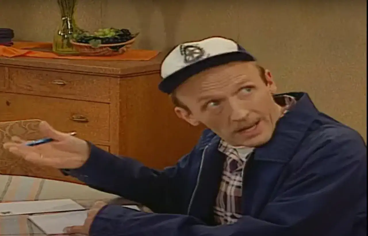 Tadeusz Norek w czapce z daszkiem i niebieskiej kurtce gestykuluje, siedząc przy stole z dokumentami.