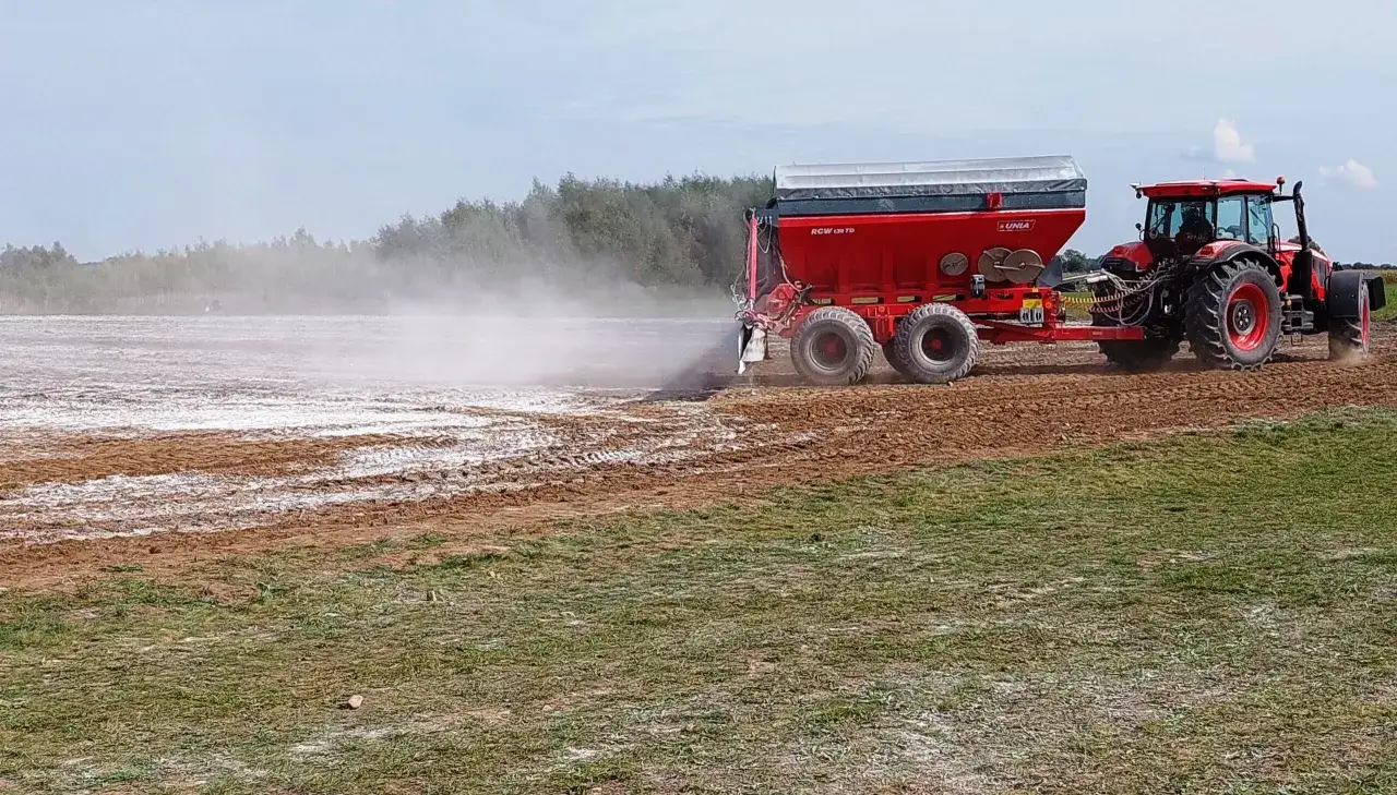 Czerwony traktor z rozsiewaczem wapna na polu. To pokazuje, co daje wapnowanie gleby – poprawia jej strukturę i żyzność.