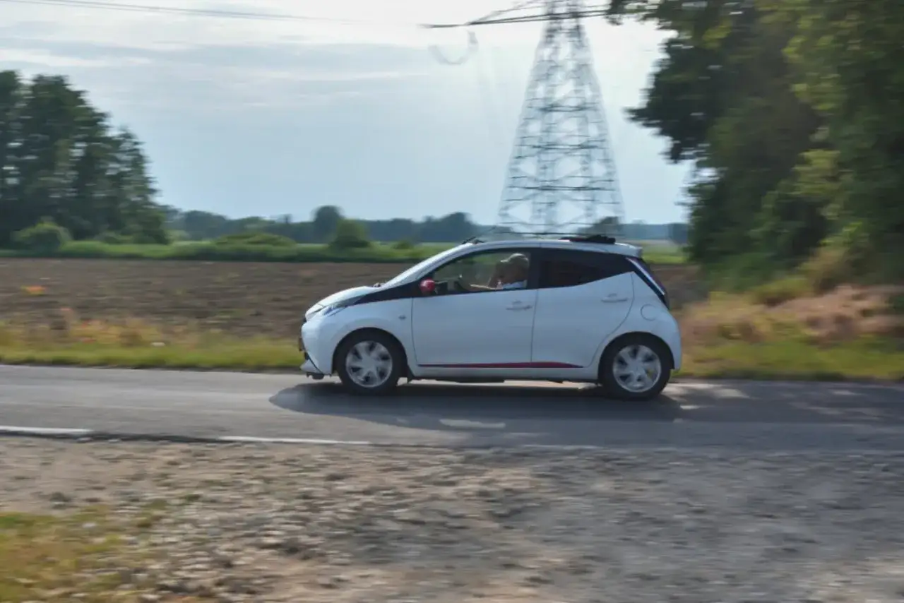 Białe auto od 14 lat z czerwonymi akcentami jedzie po wiejskiej drodze. W tle widać pola i wieżę energetyczną.