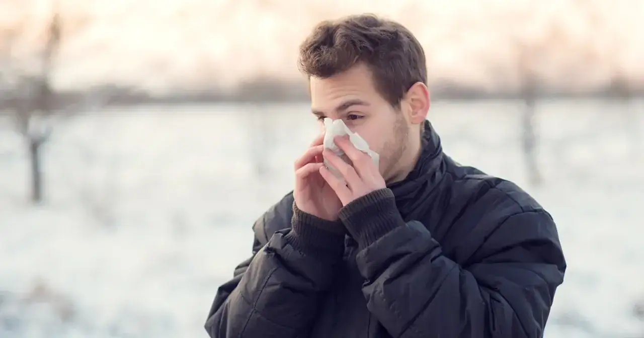 Młody mężczyzna w zimowej kurtce chusteczką wyciera nos, cierpiąc na przewlekły mokry kaszel.