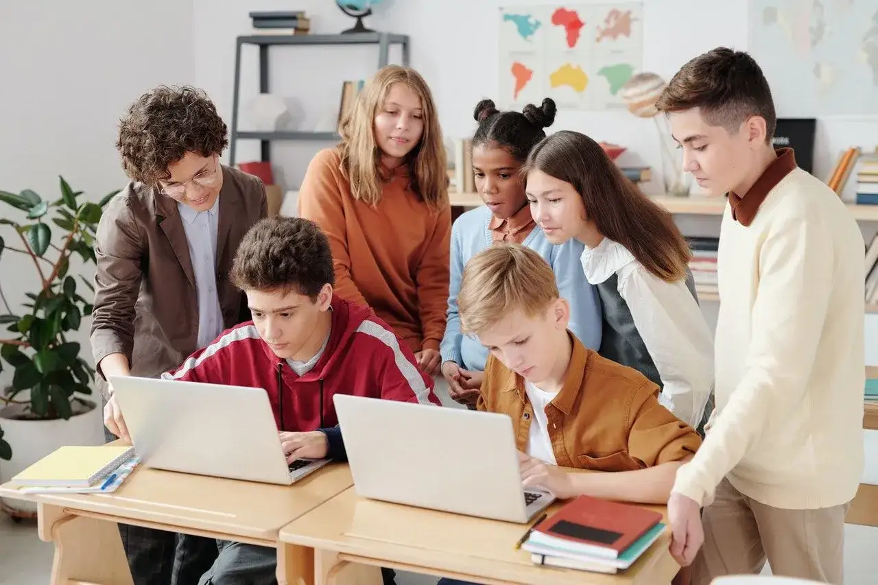 Grupa uczni&oacute;w z nauczycielem przygląda się ekranom laptop&oacute;w. Kto to? Prawdopodobnie native speakerzy uczący się czegoś nowego.