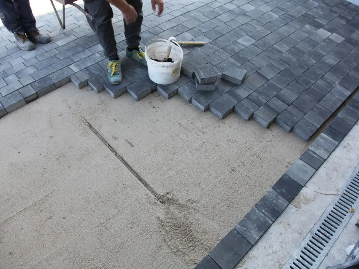 Układanie podjazdu z kostki. Pracownicy kończą montaż szarej kostki brukowej na piasku.