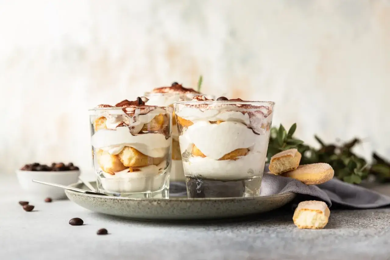 Pyszne włoskie desery tiramisu w szklankach, posypane kakao. Obok kawałki biszkopt&oacute;w i ziarna kawy.