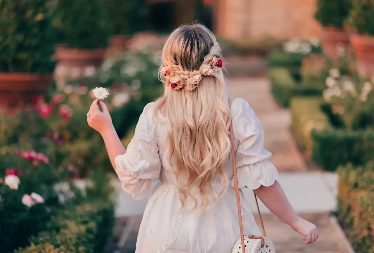Blondynka w białej sukience z bujną fryzurą boho ozdobioną kwiatami, spaceruje po ogrodzie.