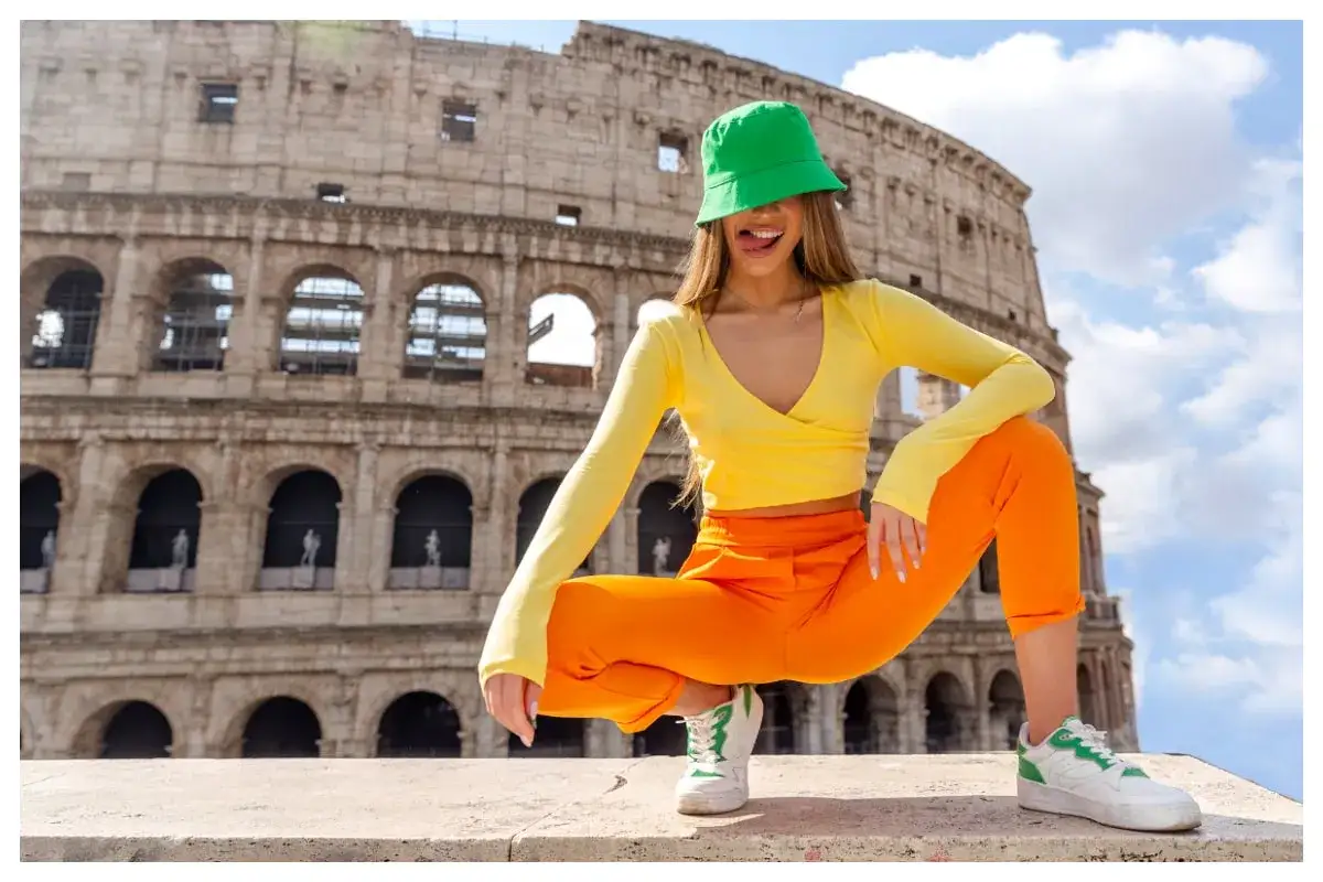 Dziewczyna w jaskrawym, pomarańczowym stroju i zielonej czapeczce kuca przed Koloseum, pokazując język.