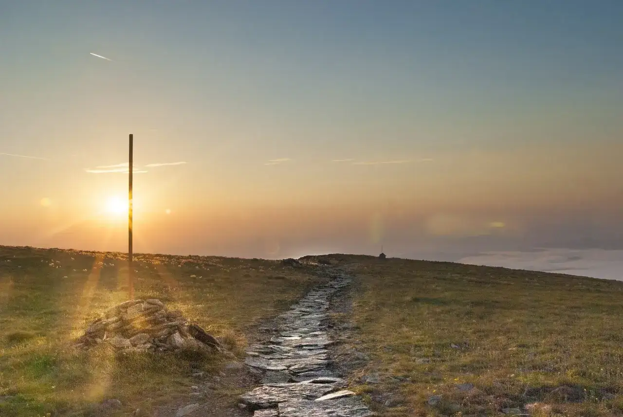 Kamienista ścieżka prowadzi przez trawiaste wzg&oacute;rza w kierunku zachodzącego słońca. W oddali widać mgłę, a na horyzoncie wieżę. To widok z hali barankowej bac&oacute;wki.