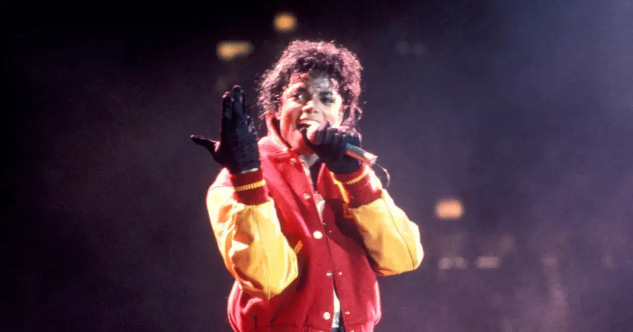 Michael Jackson na scenie, w czerwono-żółtej kurtce, śpiewa do mikrofonu. Jego gest dłonią sugeruje energię, która sprawia, że jego płyty to najważniejsze płyty w historii muzyki.