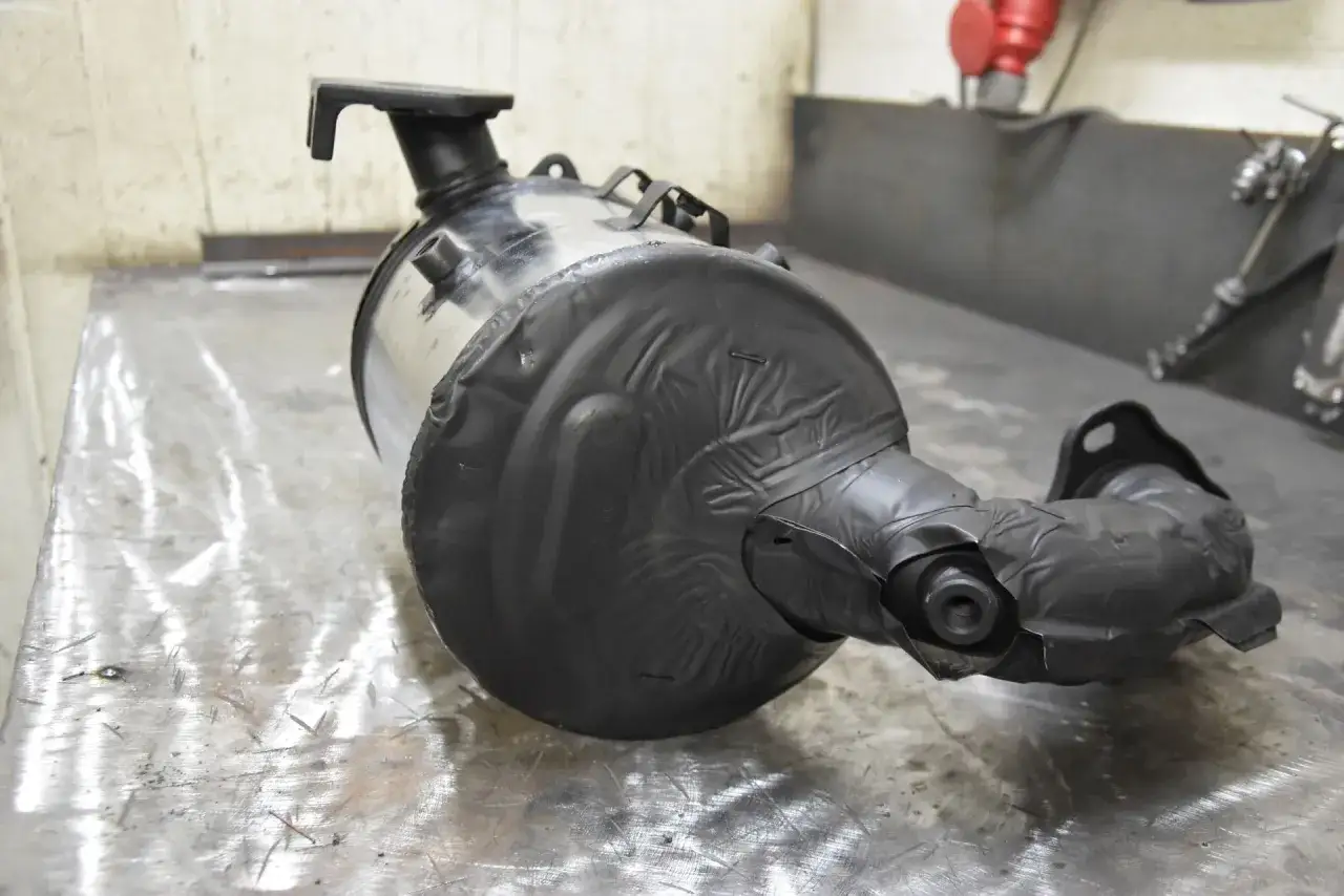 Część układu wydechowego, prawdopodobnie dpf, owinięta czarną taśmą, leży na metalowym stole.