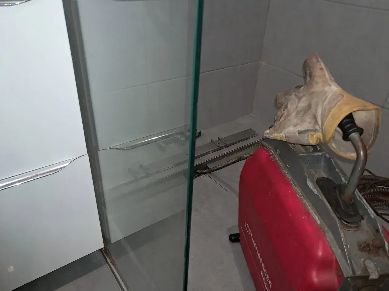 Maszyna do udrażniania rur i rękawica ochronna przy kabinie prysznicowej, gdzie widać zapchany odpływ.