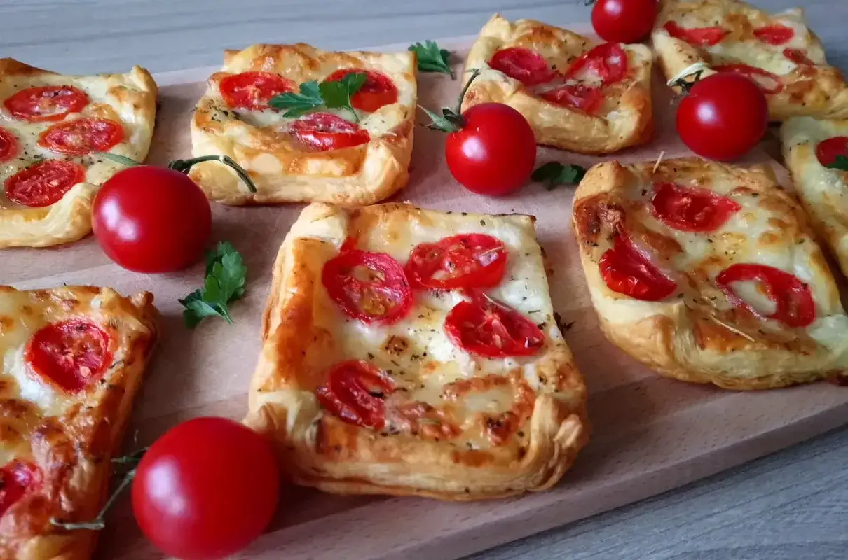 Chrupiące ciasto francuskie z serem i pomidorkami koktajlowymi, ozdobione natką pietruszki. Idealne na przekąskę.