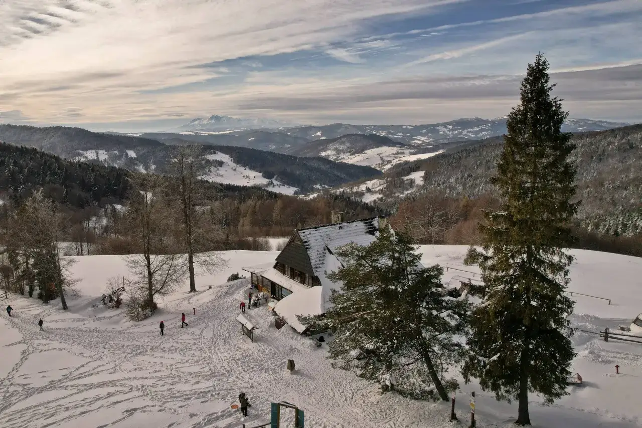Zimowy krajobraz z bac&oacute;wką nad Wierchomlą. Śnieg pokrywa wzg&oacute;rza i lasy, a w oddali widać g&oacute;ry. Kilka os&oacute;b spaceruje po zasypanym śniegiem terenie.