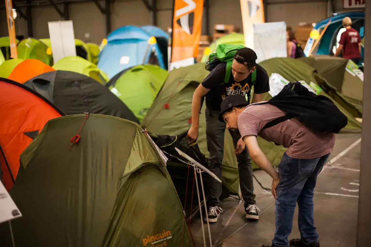 Dw&oacute;ch mężczyzn ogląda namioty, zastanawiając się, jaki namiot wybrać. W tle widać wiele kolorowych namiot&oacute;w.