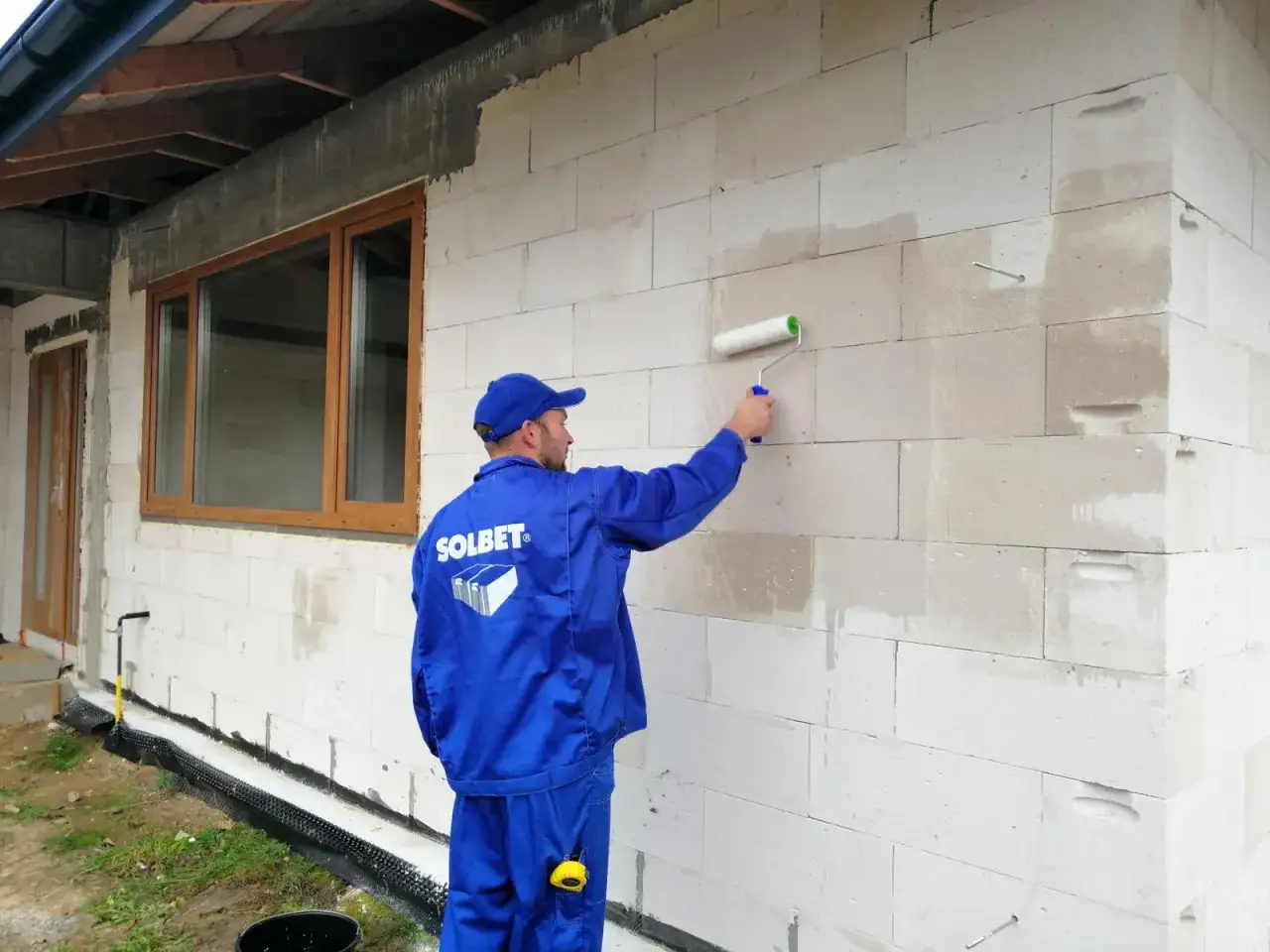 Pracownik w niebieskim kombinezonie wykonuje gruntowanie elewacji po ociepleniu, malując ścianę z bloczków betonowych wałkiem.