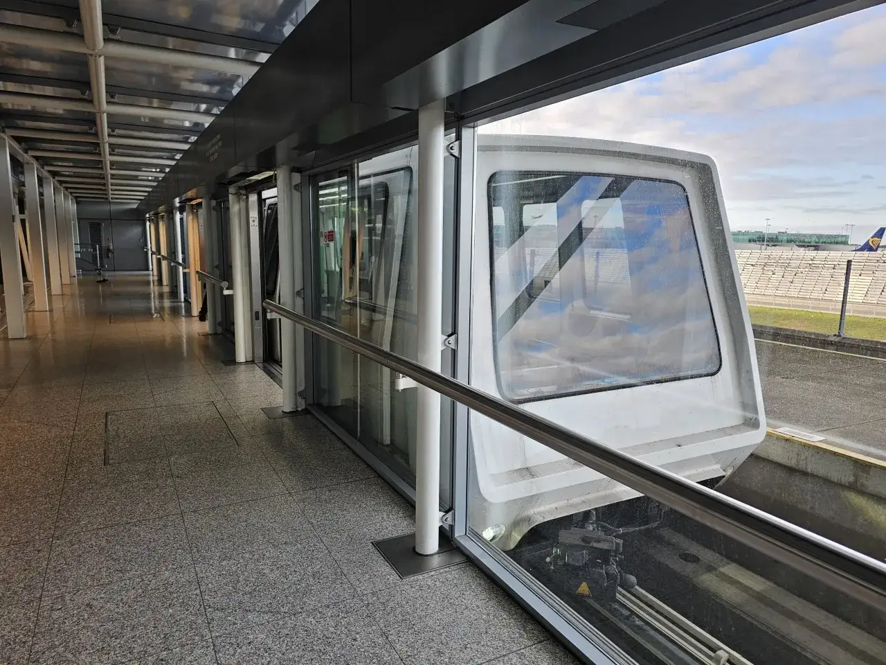 Automatyczny pociąg na lotnisku Londyn Stansted STN, z widokiem na płyty postojowe i samolot Ryanair.