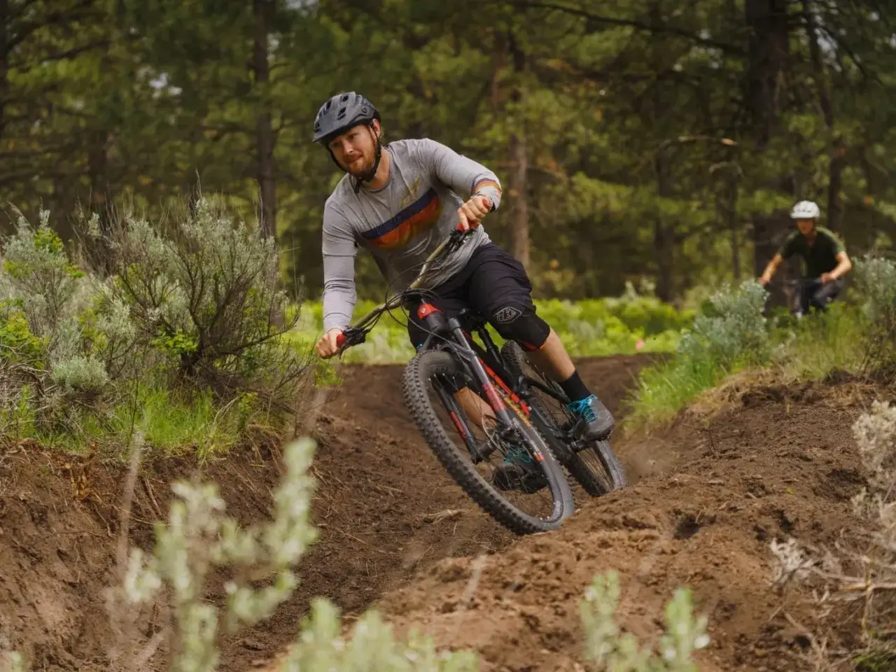 Mężczyzna w kasku zjeżdża na rowerze po leśnym szlaku. W tle inny rowerzysta.