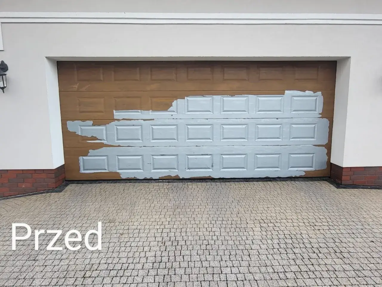 Przed wymianą okleiny na bramie garażowej. Drewniana powierzchnia jest częściowo pokryta jasnoszarą farbą, kt&oacute;ra łuszczy się, odsłaniając pierwotny kolor.