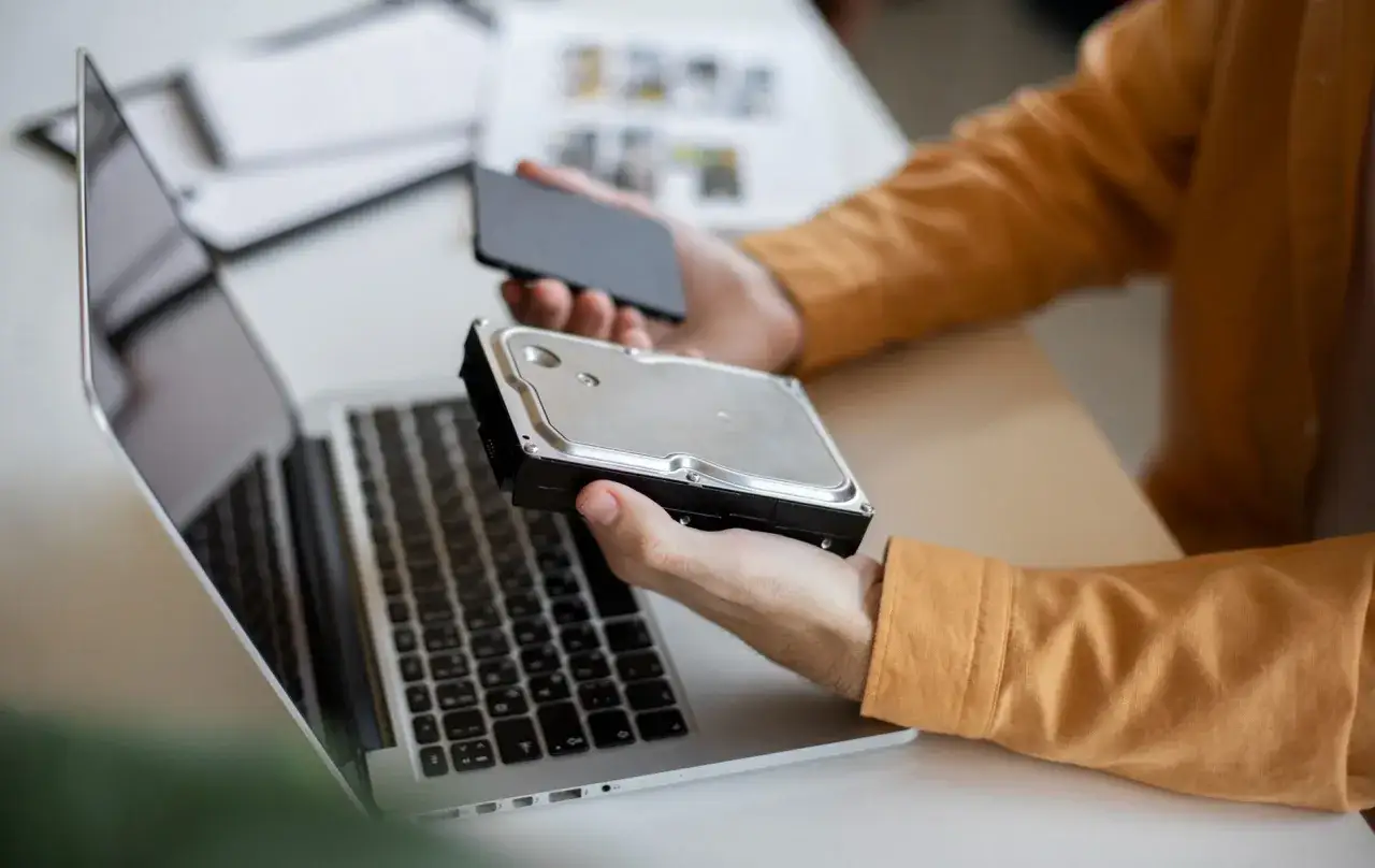 Osoba w pomarańczowej koszuli przygotowuje klonowanie dysku twardego, trzymając go obok laptopa i smartfona.