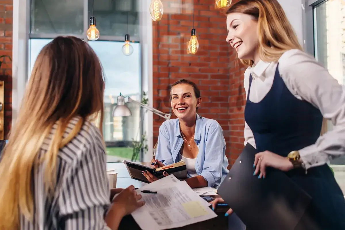 Trzy kobiety rozmawiają w biurze. Jedna z nich, w jasnej koszuli, uśmiecha się, trzymając notatnik. To idealny moment, by powiedzieć dziewczynie, co powiedzieć dziewczynie żeby czuła się wyjątkowa.