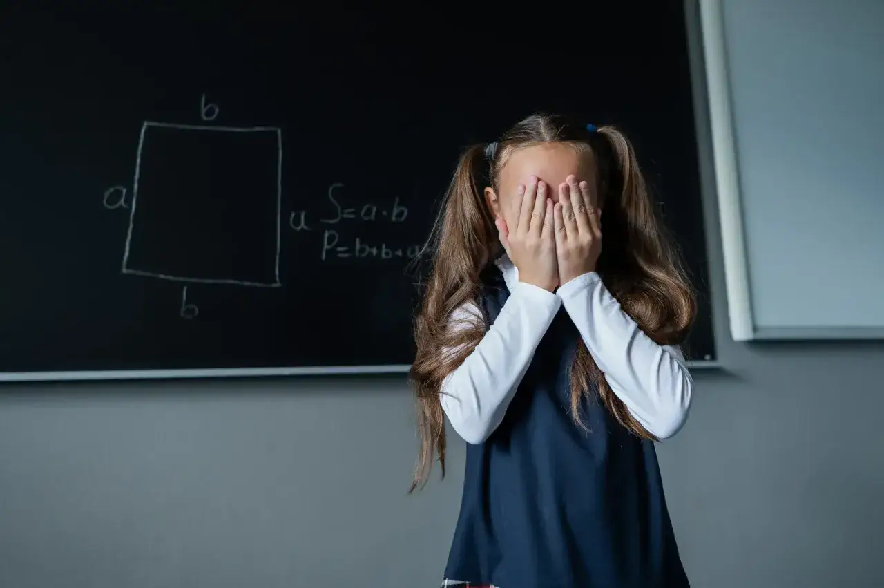 Dziewczynka z opuszczonymi rękami na twarzy, przed tablicą z wzorami matematycznymi, wyraźnie przeżywa problemy z matematyką.