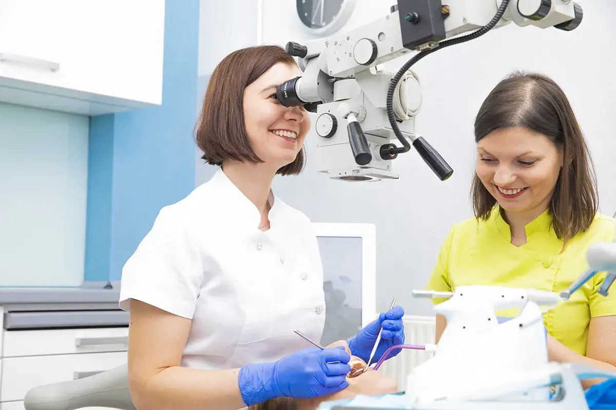 Uśmiechnięta dentystka w białym fartuchu bada ząb pacjenta przez mikroskop. Obok stoi asystentka. To, co widzi dentysta, jest kluczowe dla precyzyjnego leczenia.