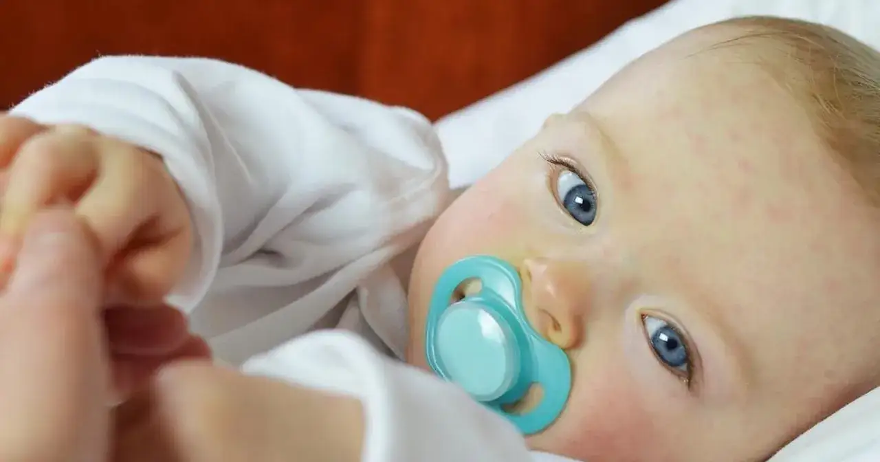 Niemowlę z niebieskimi oczami i smoczkiem ma na policzkach zaczerwienienia, kt&oacute;re mogą wskazywać na skazę białkową u niemowlaka.