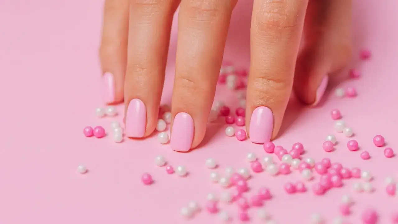 Delikatny różowy manicure hybrydowy na zadbanych paznokciach, otoczony słodkimi kuleczkami w odcieniach różu i bieli.
