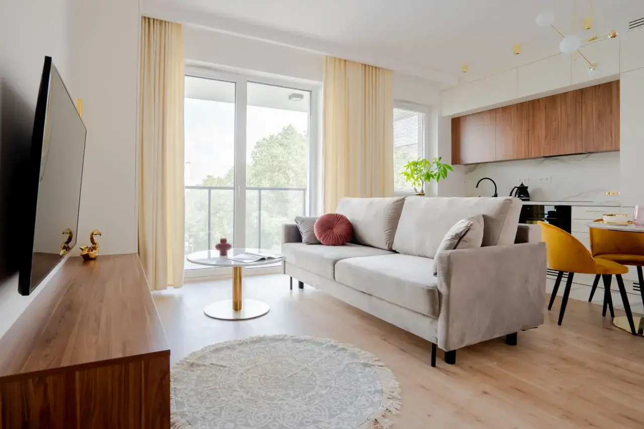 Jasne, nowoczesne mieszkanie 40m2 rzut. Salon z beżową kanapą, stolikiem kawowym i telewizorem. W tle kuchnia z drewnianymi frontami i ż&oacute;łte krzesła.