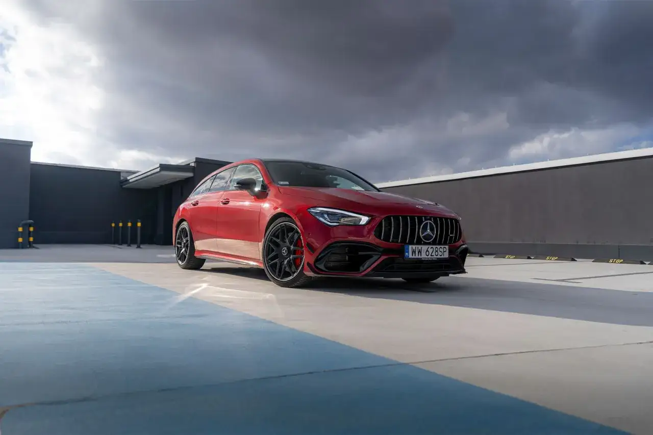 Czerwony Mercedes CLA 45 AMG na parkingu pod pochmurnym niebem. Agresywny grill i sportowe felgi podkreślają jego moc.