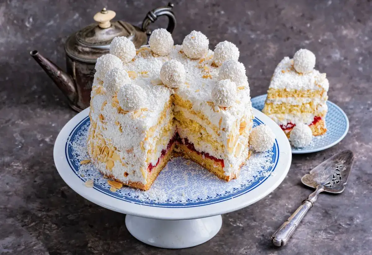 Pyszne ciasto rafaello na biszkopcie, udekorowane wi&oacute;rkami kokosowymi i kulkami Rafaello. Obok kawałek ciasta na talerzyku.