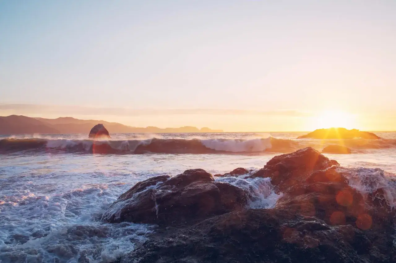 Zach&oacute;d słońca nad morzem. Fale rozbijają się o skały, tworząc pianę. W oddali widać g&oacute;ry.