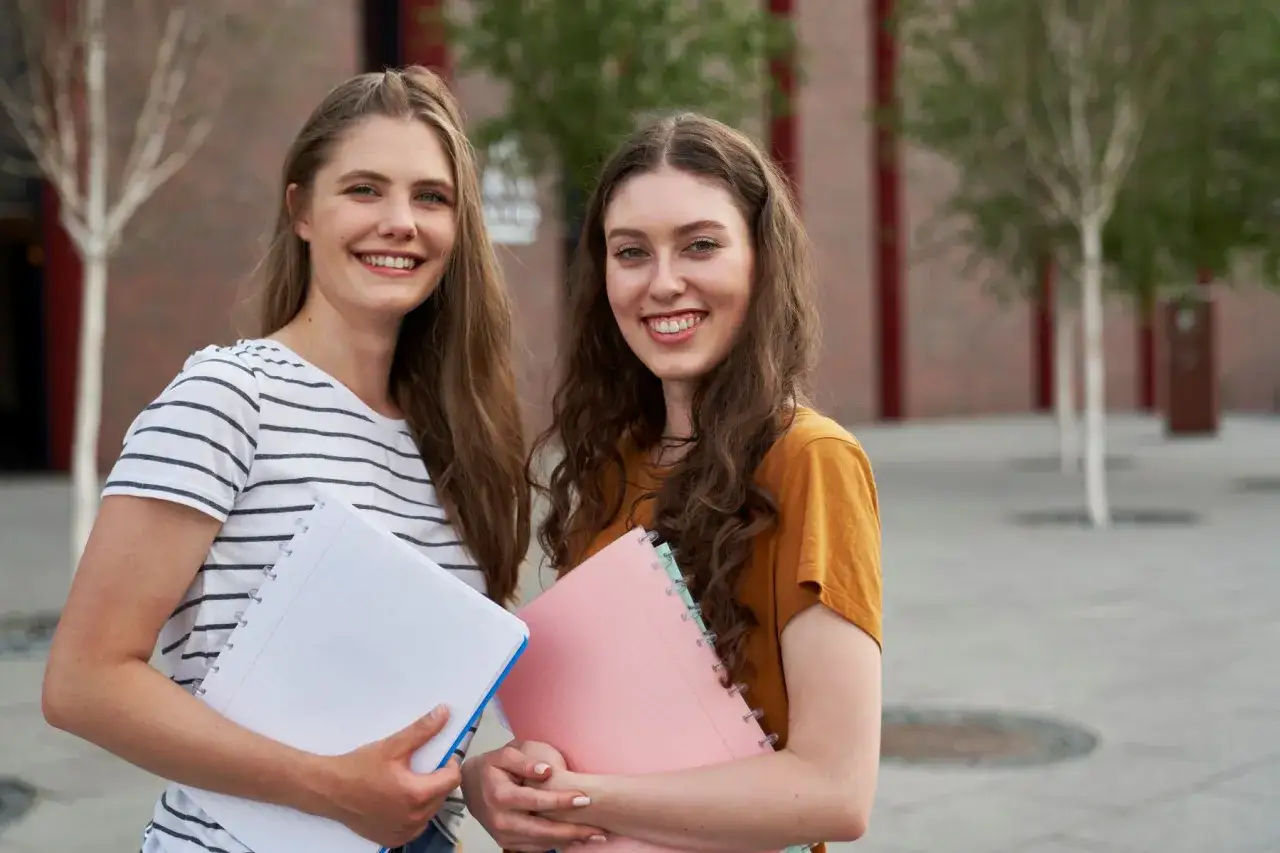 Dwie uśmiechnięte studentki z notatnikami. Czy na studium trzeba mieć maturę? Tak, to podstawa!