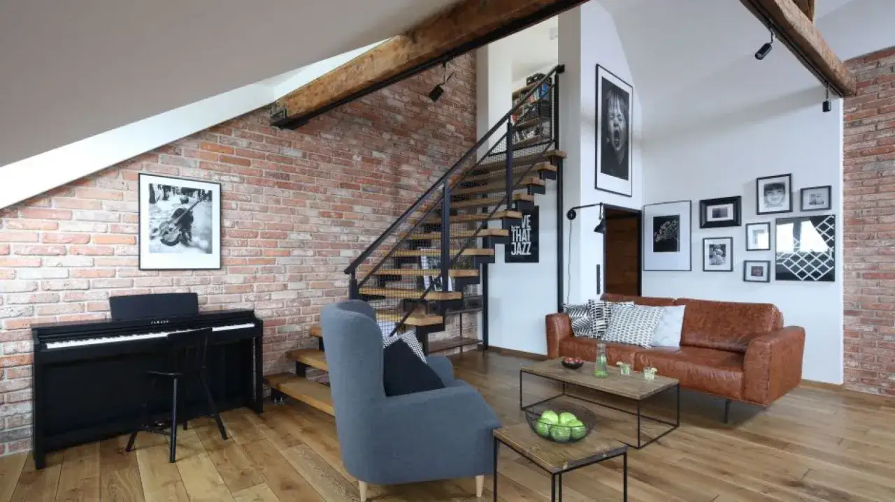 Nowoczesny salon z ceglaną ścianą, schodami, pianinem i wygodną kanapą. Oświetlenie ściany z cegły podkreśla jej fakturę.