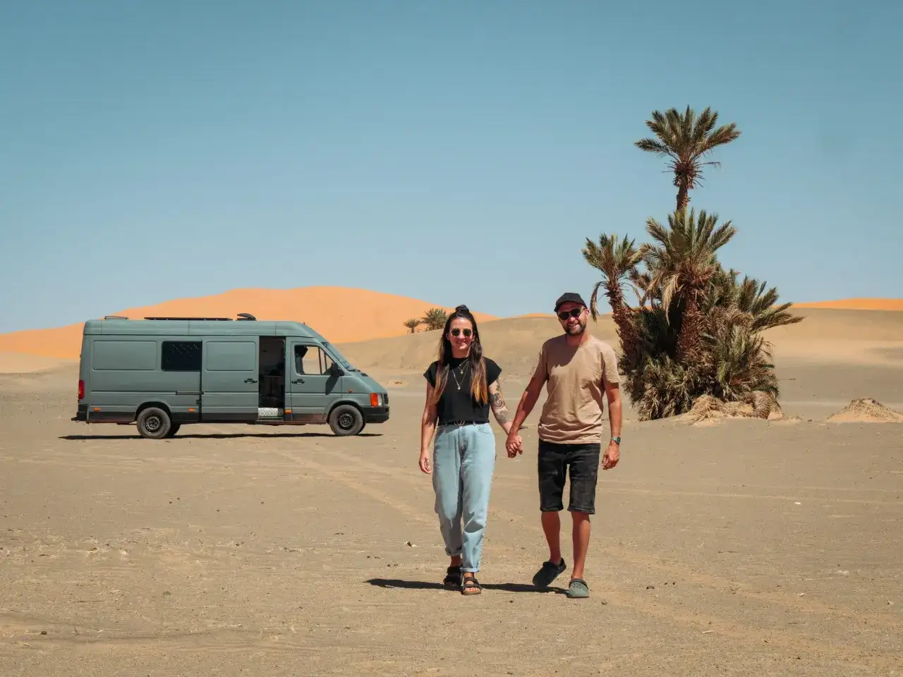 Para wędruje przez pustynię obok swojego szarego vana mieszkalnego, z palmami w tle.
