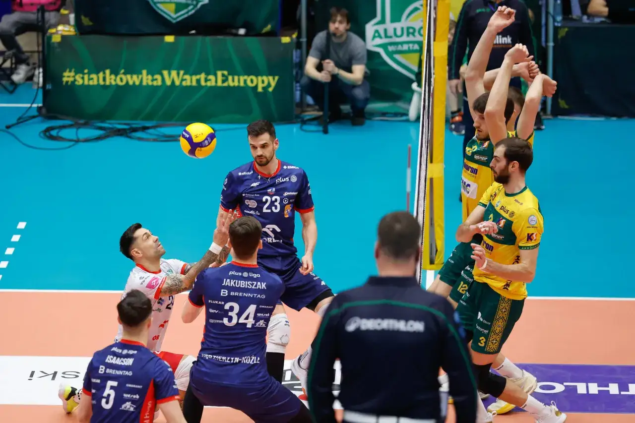 Siatkarze walczą o piłkę w meczu Plus Ligi. Na boisku widać zawodników w niebieskich i żółtych strojach.