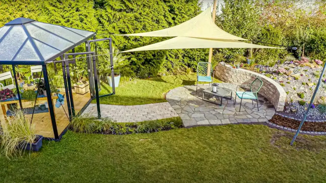 Jak zamontować żagiel na tarasie? W ogrodzie z szklarnią, kamiennym patio i skalniakiem, dwa beżowe żagle tworzą zacienione miejsce do wypoczynku.