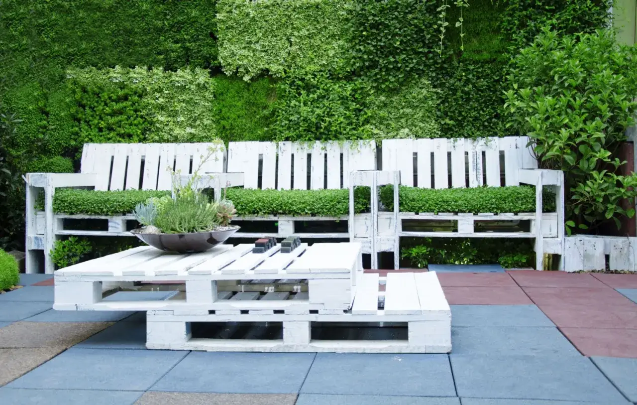 Meble ogrodowe z palet, idealne rozwiązanie, co można zrobić ze starych desek. Biała ławka i stolik z roślinnością.