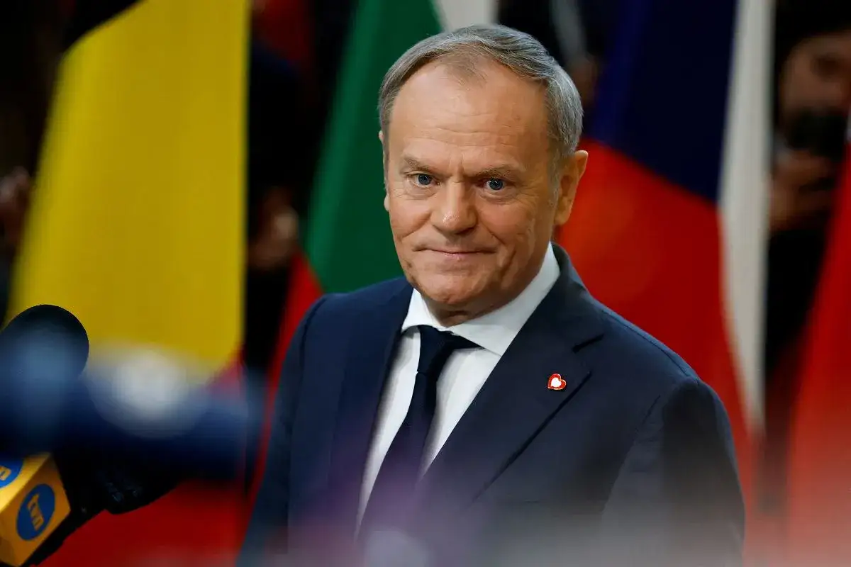 Donald Tusk, w garniturze, z flagami w tle. Zastanawiamy się, co zrobił Tusk dla Polski, gdy patrzy w obiektyw.