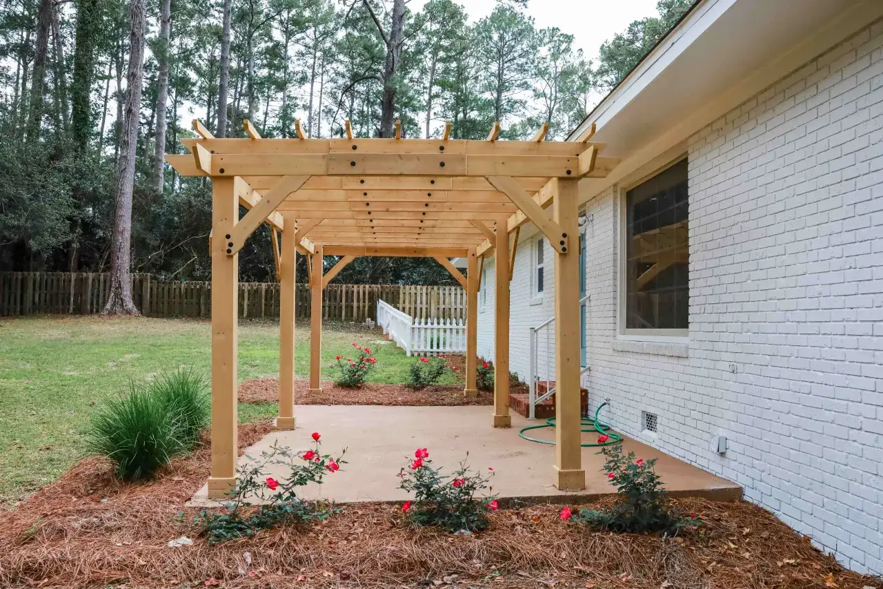 Drewniana pergola przy białym domu, z czerwonymi różami i zieloną trawą. Pokazano, jak zakotwić pergolę.