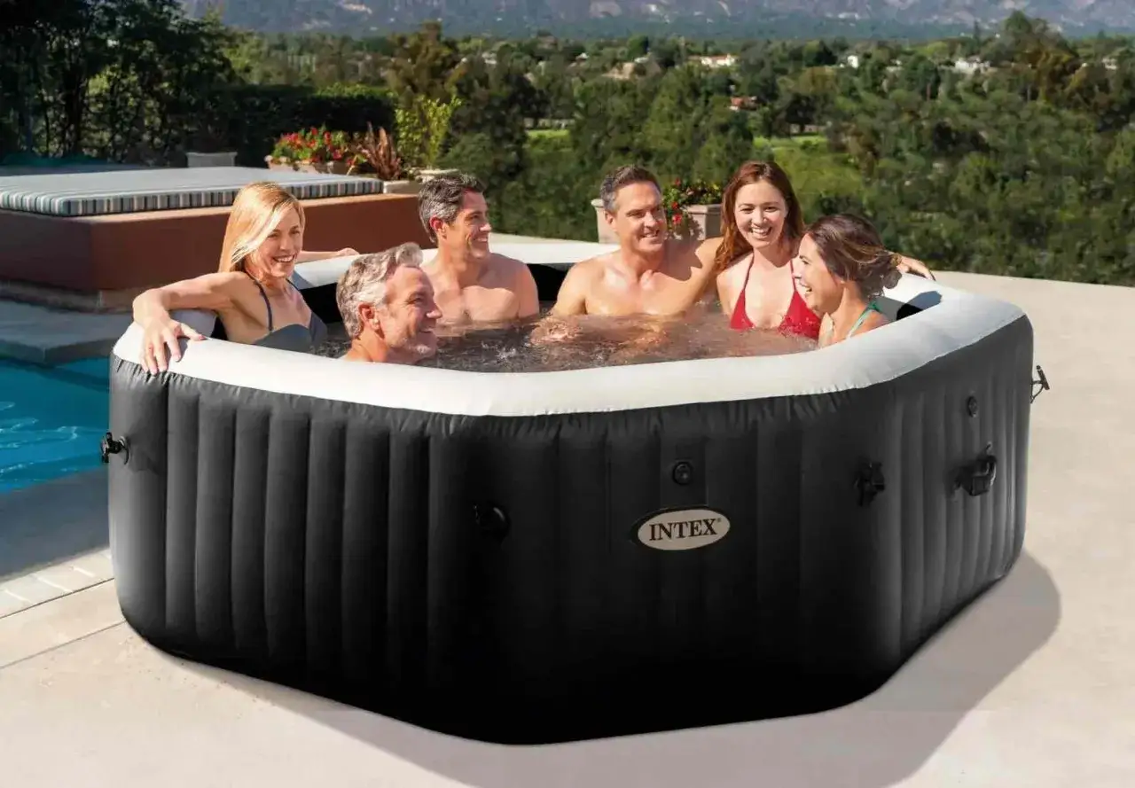 Sześć os&oacute;b śmieje się i relaksuje w dmuchanym jacuzzi Intex Pure Spa na tarasie z widokiem na wzg&oacute;rza.
