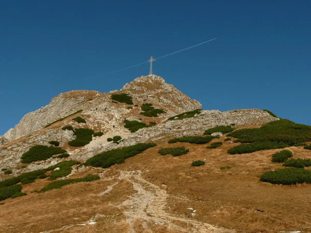 Kamienista ścieżka prowadzi na skaliste szczyty w Tatrach, gdzie na jednym z nich widać krzyż. Na błękitnym niebie zostawia smugę samolot.