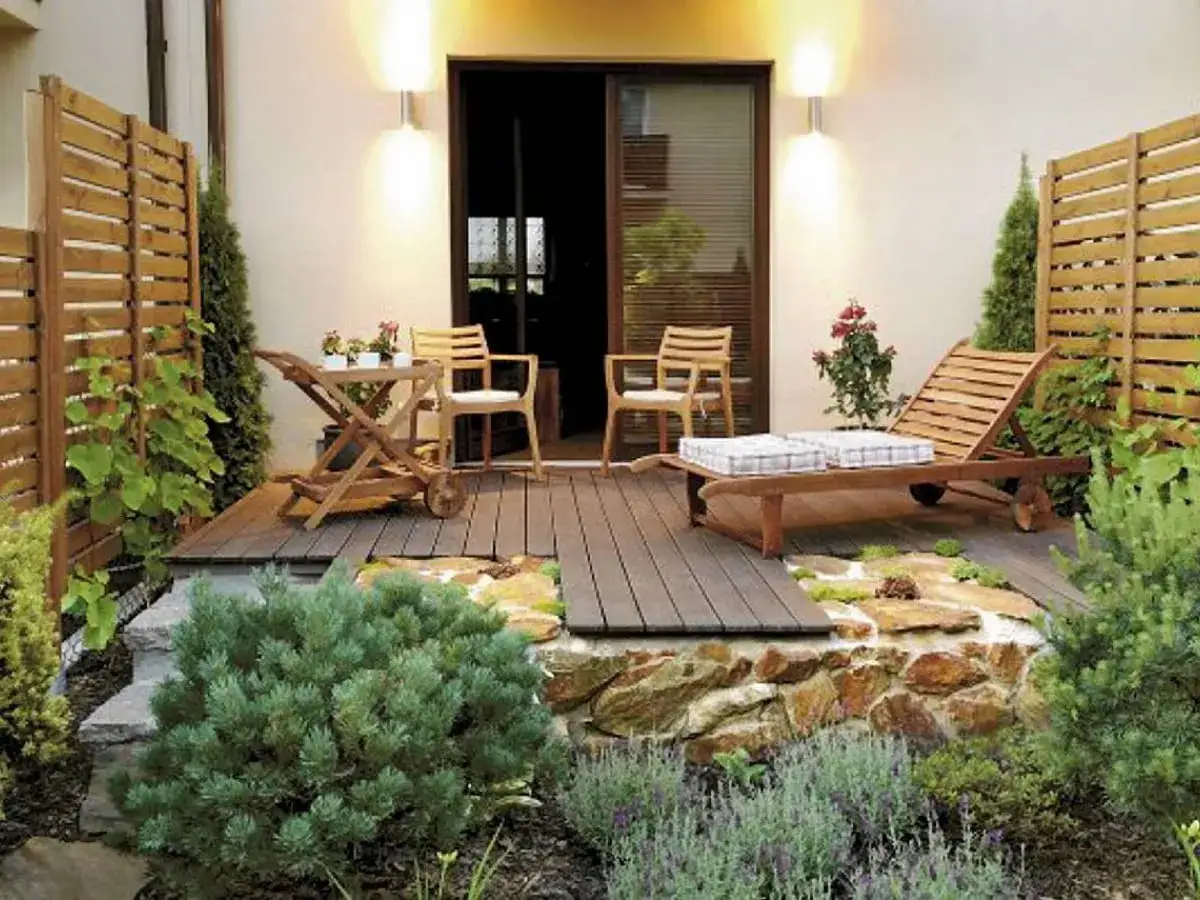 Piękny mały ogród z drewnianym tarasem, leżakiem, stolikiem i krzesłami. Kamienna rabata z roślinnością dodaje uroku.