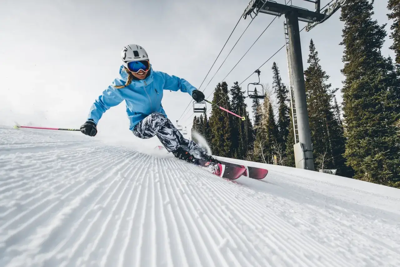Uśmiechnięta narciarka w niebieskiej kurtce i wzorzystych spodniach zjeżdża po stoku, pokazując, jak jeździć na nartach. W tle widać wyciąg krzesełkowy i drzewa.