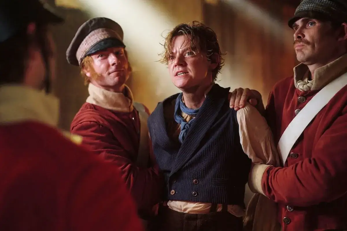 Młody mężczyzna, jak artful dodger, w ciemnej kamizelce i z niebieską bandaną, otoczony przez żołnierzy w czerwonych kurtkach.